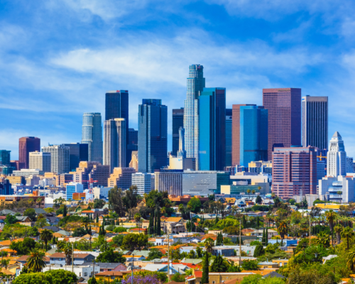 The downtown Los Angeles skyline under a bright blue sky, featuring modern skyscrapers and colorful mid-rise buildings surrounded by residential neighborhoods and palm trees. The image represents how eminent domain and property tax laws impact property owners across the diverse Los Angeles landscape.