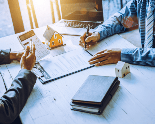 Two individuals reviewing and signing a contract at a desk with a calculator, laptop, and small house model, representing property valuation, real estate transactions, and legal agreements in eminent domain cases.