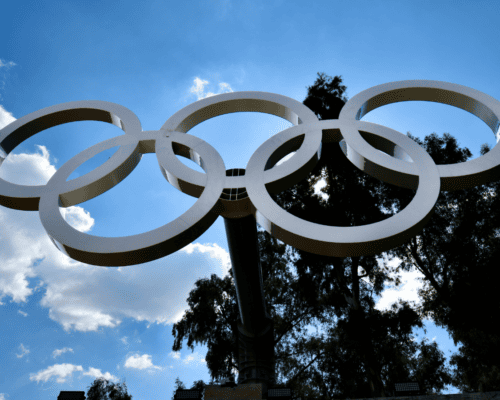 A large Olympic rings sculpture set against the Los Angeles skyline, symbolizing upcoming 2028 Olympic infrastructure projects that may impact nearby properties through eminent domain and public development.